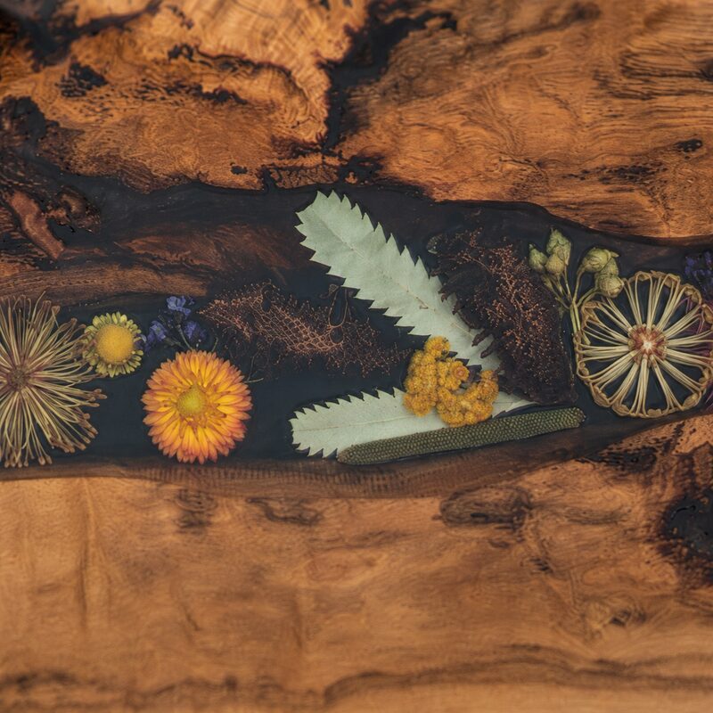 Close-up of preserved Australian wildflowers and native foliage encased in smooth dark resin beside richly figured timber