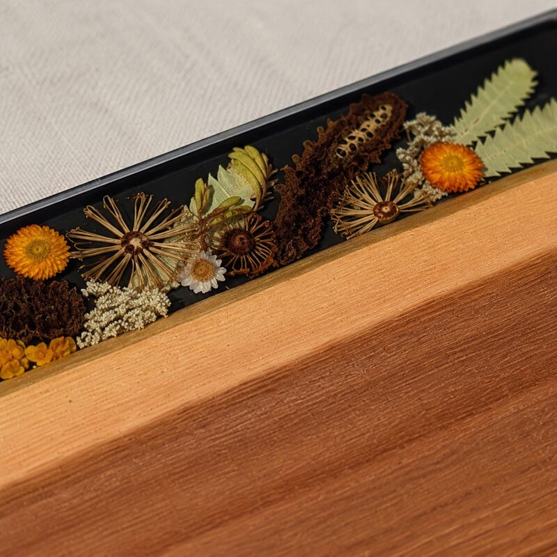 Close-up of preserved Australian flowers, fern leaves and seedpods beneath smooth dark resin beside two-tone timber.