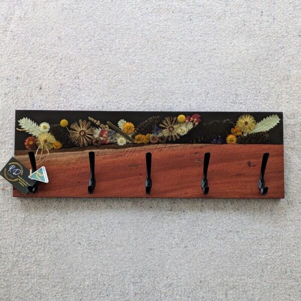 The top portion of the rack showcases a stunning, hand-poured resin inlay embedded with real, carefully preserved Australian wildflowers and foliage. Every plant and flower is thoughtfully arranged and sealed in crystal-clear resin, capturing the delicate beauty of nature in timeless detail. Below, the warm tones and unique grain of the locally sourced timber add rustic charm, creating a perfect balance between natural texture and glossy sophistication.